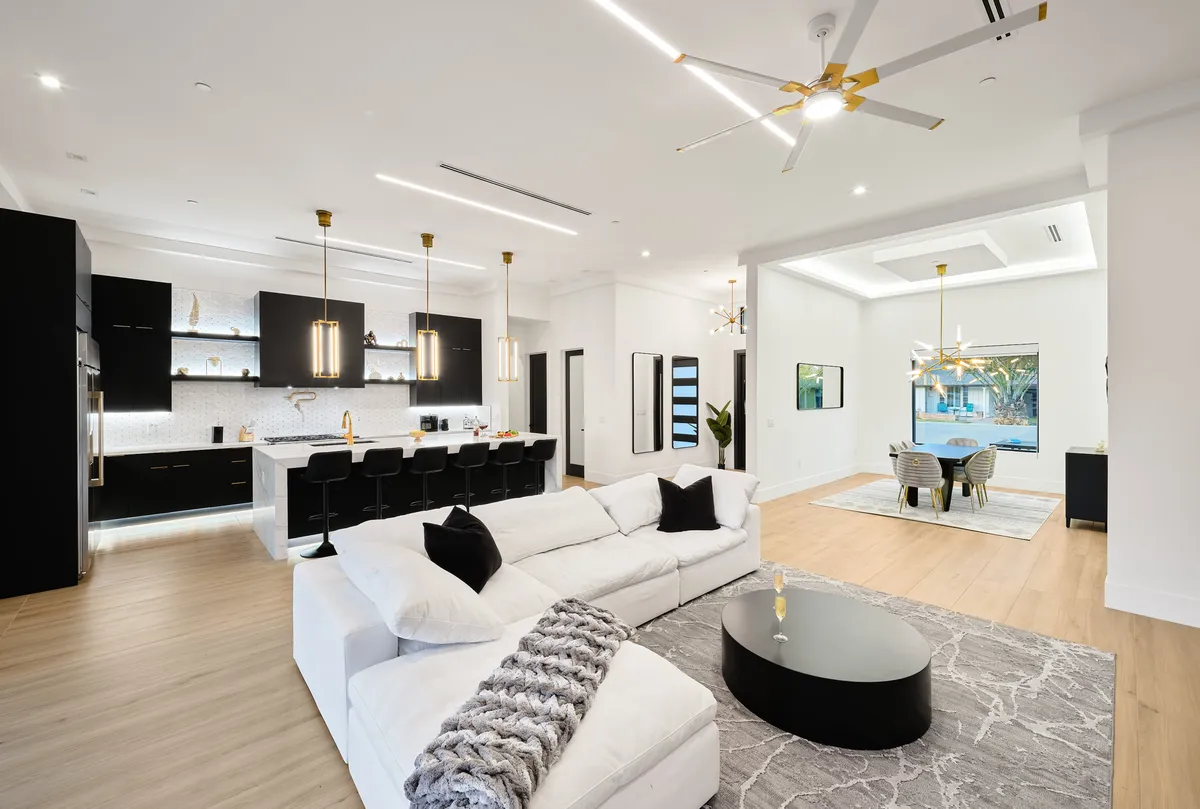A sweeping open-concept space featuring a modern sectional sofa in the living area, a sleek kitchen island with bar seating, and an elegant dining table. The styling is contemporary, with plush furnishings, marble finishes, and designer lighting that unify the space. Artwork and greenery add warmth and sophistication.