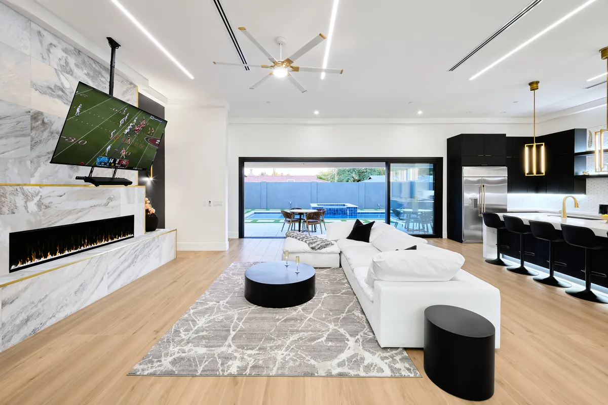 A stylish, contemporary living area with a marble fireplace and large TV, white sectional sofa, black accent table, and sliding glass doors opening to the outdoor space. The space is bright, inviting, and suitable for family relaxation or group gatherings.