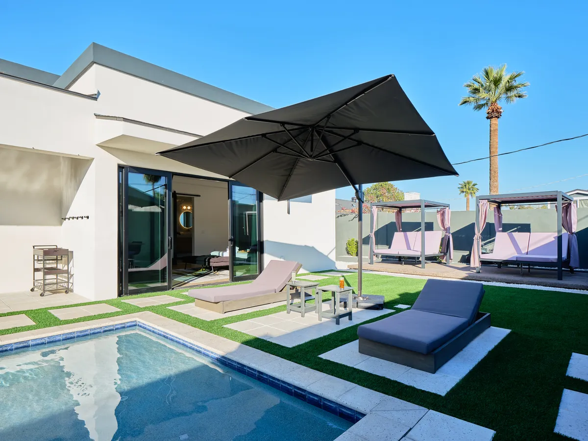 A sparkling swimming pool serves as the centerpiece, surrounded by modern lounge chairs, large umbrellas, and outdoor cabanas. Lush green lawn and vibrant landscaping enhance the inviting, resort-like feel. The space is bathed in warm sunset light, hinting at both daytime fun and evening relaxation.