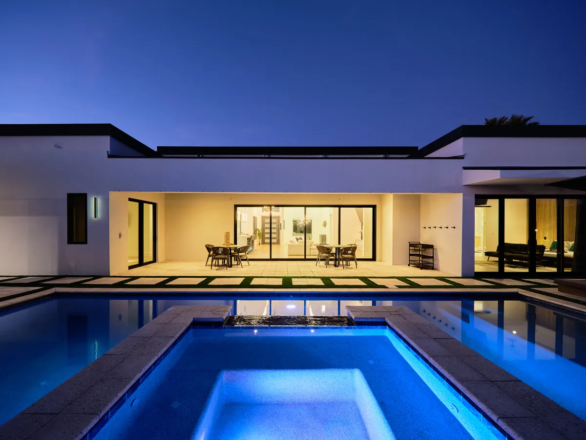A sparkling swimming pool sits adjacent to an elegant outdoor dining area, with large glass doors opening up to the space. Subtle lighting suggests the area is ready for evening gatherings, while the setup hints at al fresco meals and special celebrations by the water.