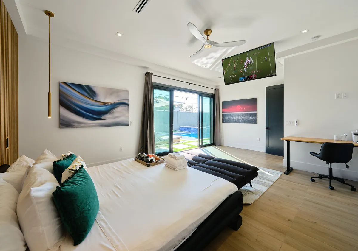 This image features a spacious bedroom with a bed made up with plush pillows and bedding, a large window offering an outdoor view, a wall-mounted television, tasteful artwork, and a wooden desk with a chair. The setting is modern, comfortable, and designed for both relaxation and productivity.