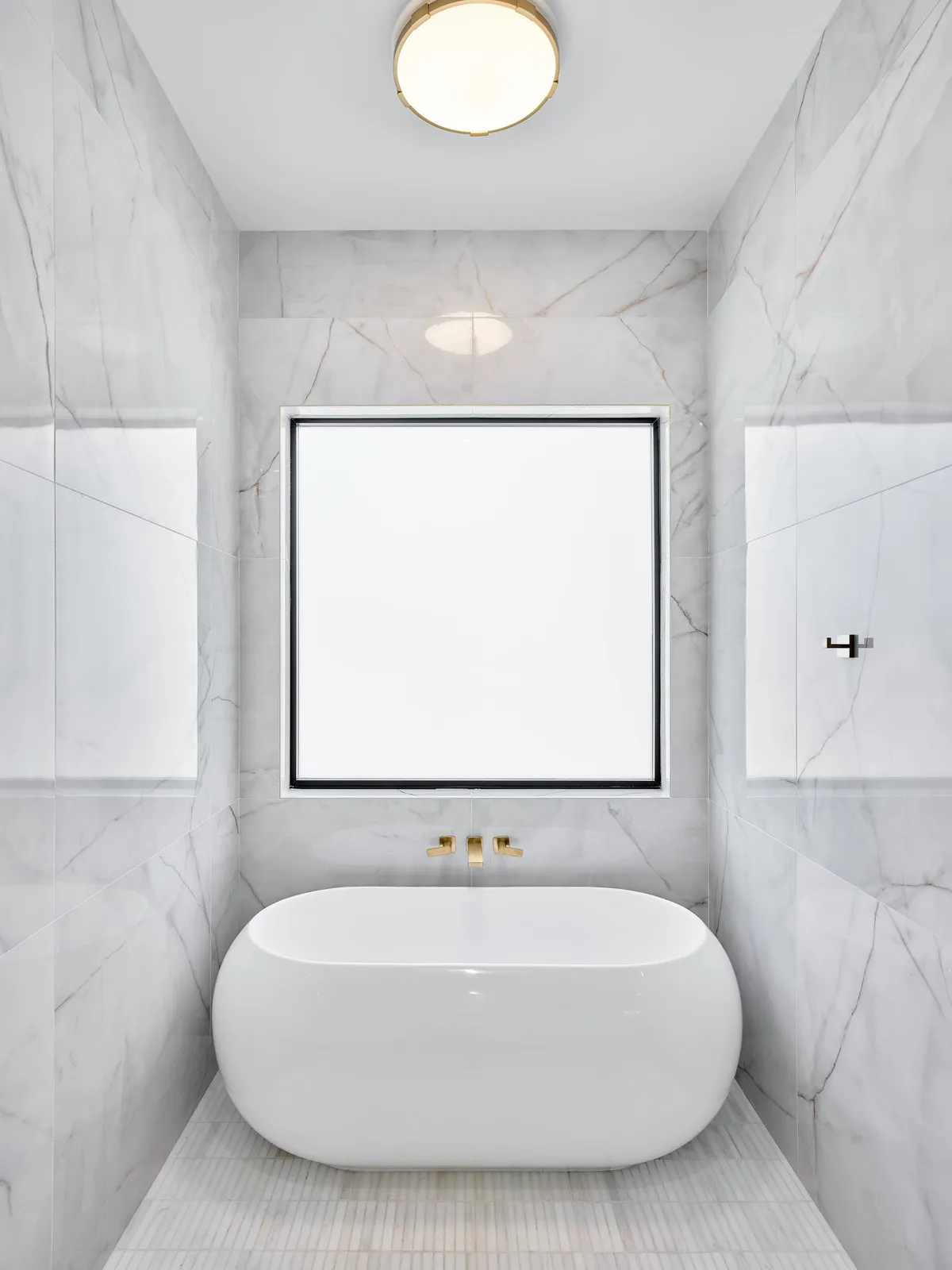 A luxurious bathroom with marble walls, a freestanding soaking tub positioned near a large window, gold fixtures, and tranquil natural light. The space radiates spa-like calm and indulgence, perfect for unwinding after a busy day.