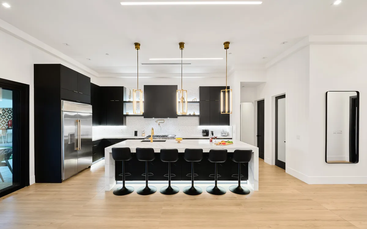 A close-up view of a high-end kitchen showcasing a large island with pendant lighting, sleek black cabinetry, and top-of-the-line stainless steel appliances. The mood is sophisticated, and the finishes include marble countertops and modern fixtures.