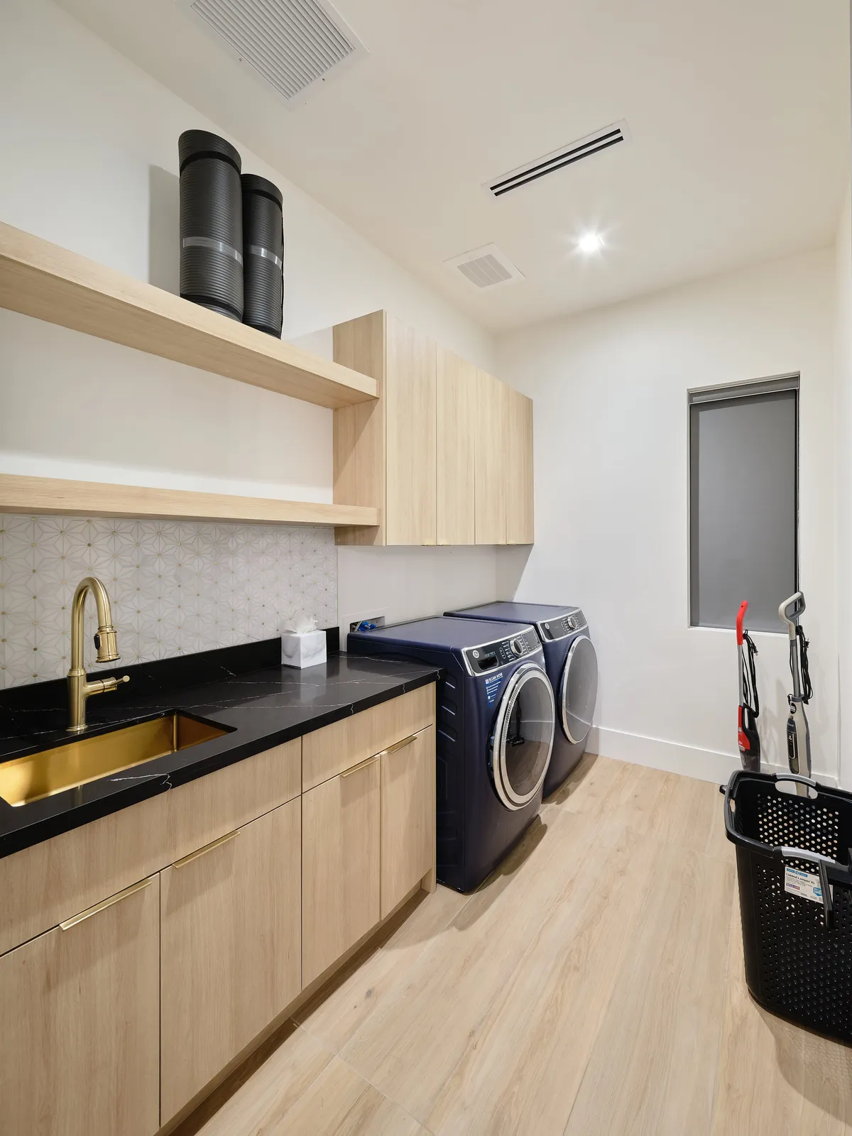 A functional, modern laundry room featuring a washing machine and dryer, gold faucet, black countertop, and wooden cabinets. The space is organized, clean, and showcases the convenience of in-home household management.
