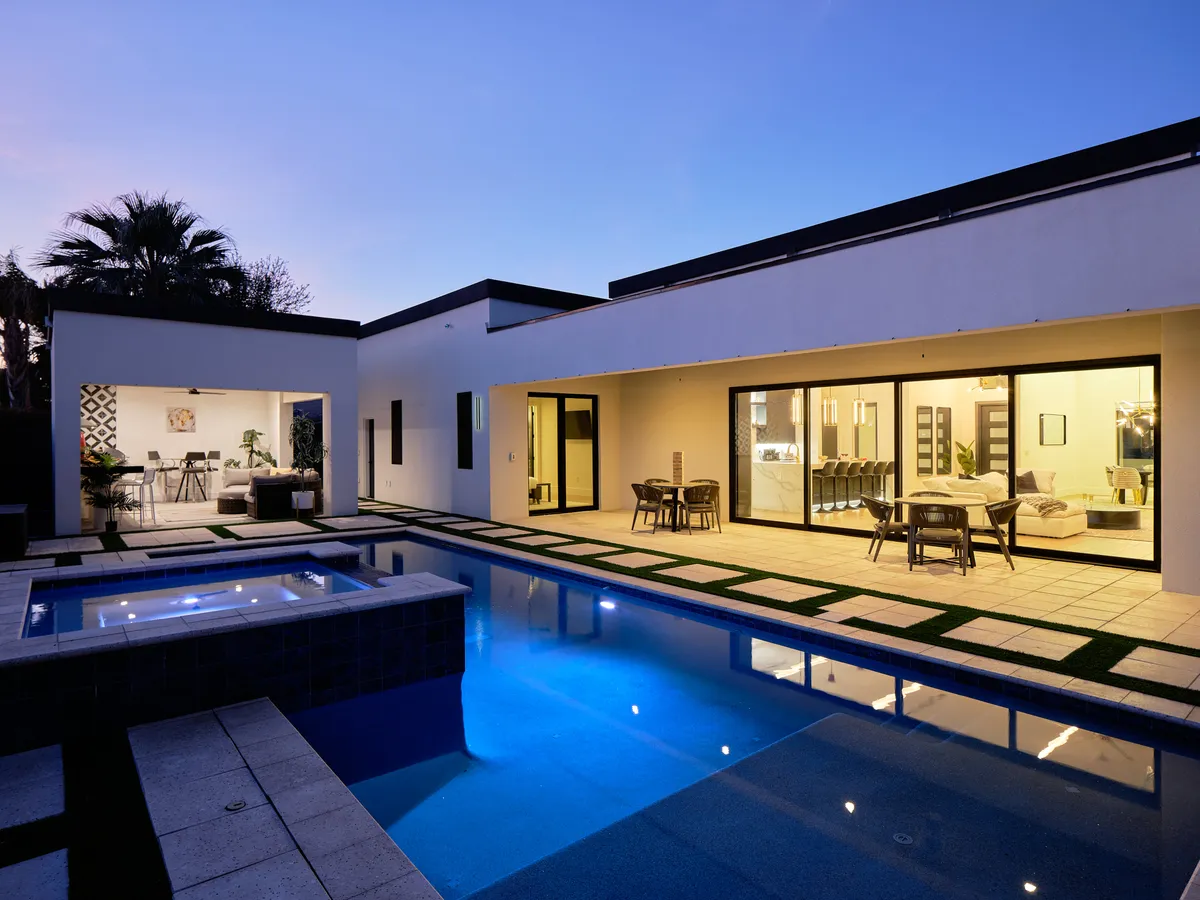 The image showcases a luxurious backyard oasis at dusk, featuring a sparkling heated swimming pool surrounded by stylish loungers and an inviting outdoor seating area. Large glass windows offer a glimpse of the modern, light-filled interior, while the scene is bathed in the warm glow of sunset. The atmosphere is serene yet vibrant, perfect for evening swims, al fresco dinners, and unwinding under the Arizona sky.